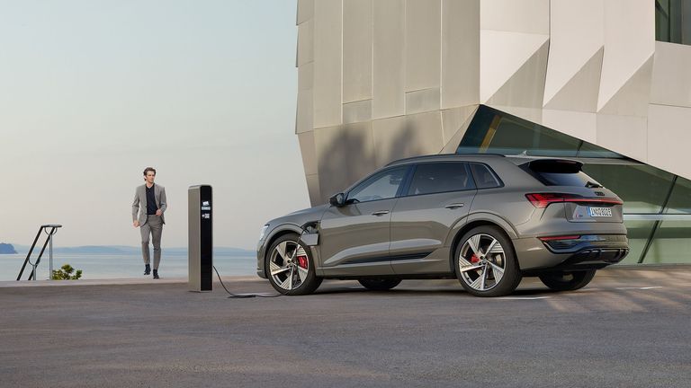 „Chronos Grey Metallic“ spalvos „Audi Q8 e-tron“, prijungtas prie įkrovimo stotelės priešais pastatą.